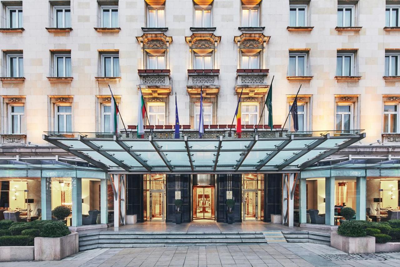 Sofia Balkan Palace Exterior photo The photo shows the exterior of a luxury hotel. The building features a classic architectural style with a light-colored facade, adorned with decorative elements such as balconies and window frames. Above the entrance, there is a stylish glass canopy